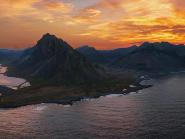 Aerial view of a jagged, dark mountain by the sea, with a long sand spit enclosing a calm lagoon, under a vibrant orange sunset sky.