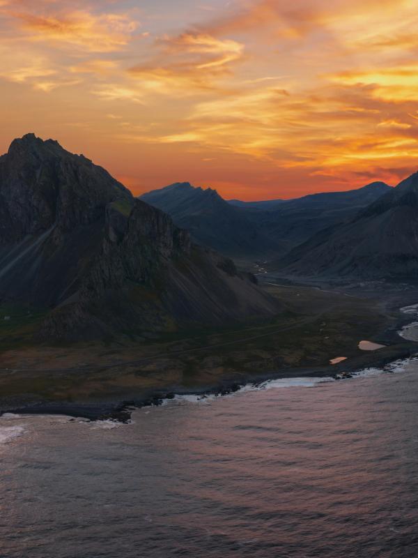 Jagged mountains meet a dramatic coastline and calm bay under a vibrant orange sunset.