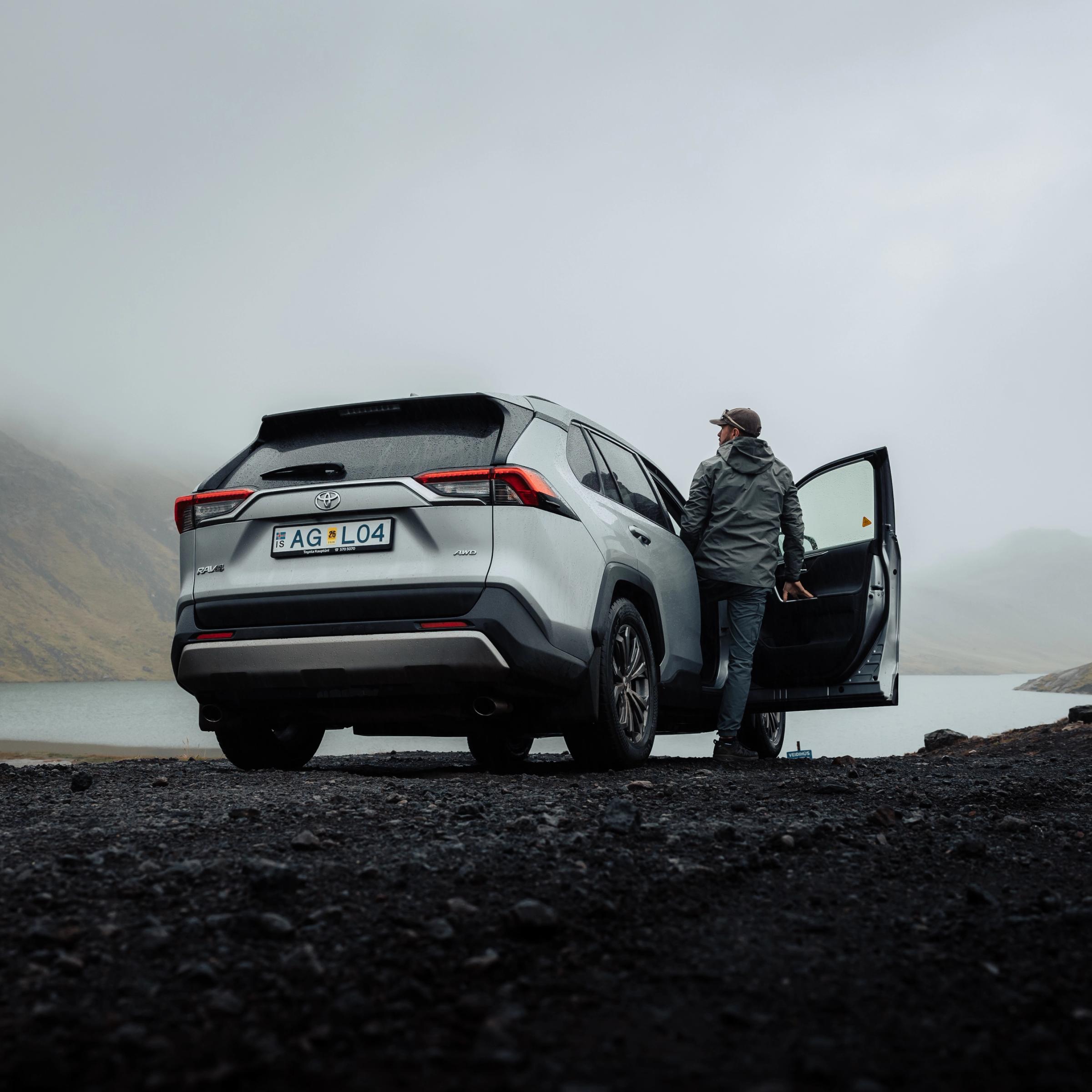 Una persona está junto a un SUV plateado con la puerta abierta sobre un terreno oscuro, contemplando un lago brumoso y unas montañas.