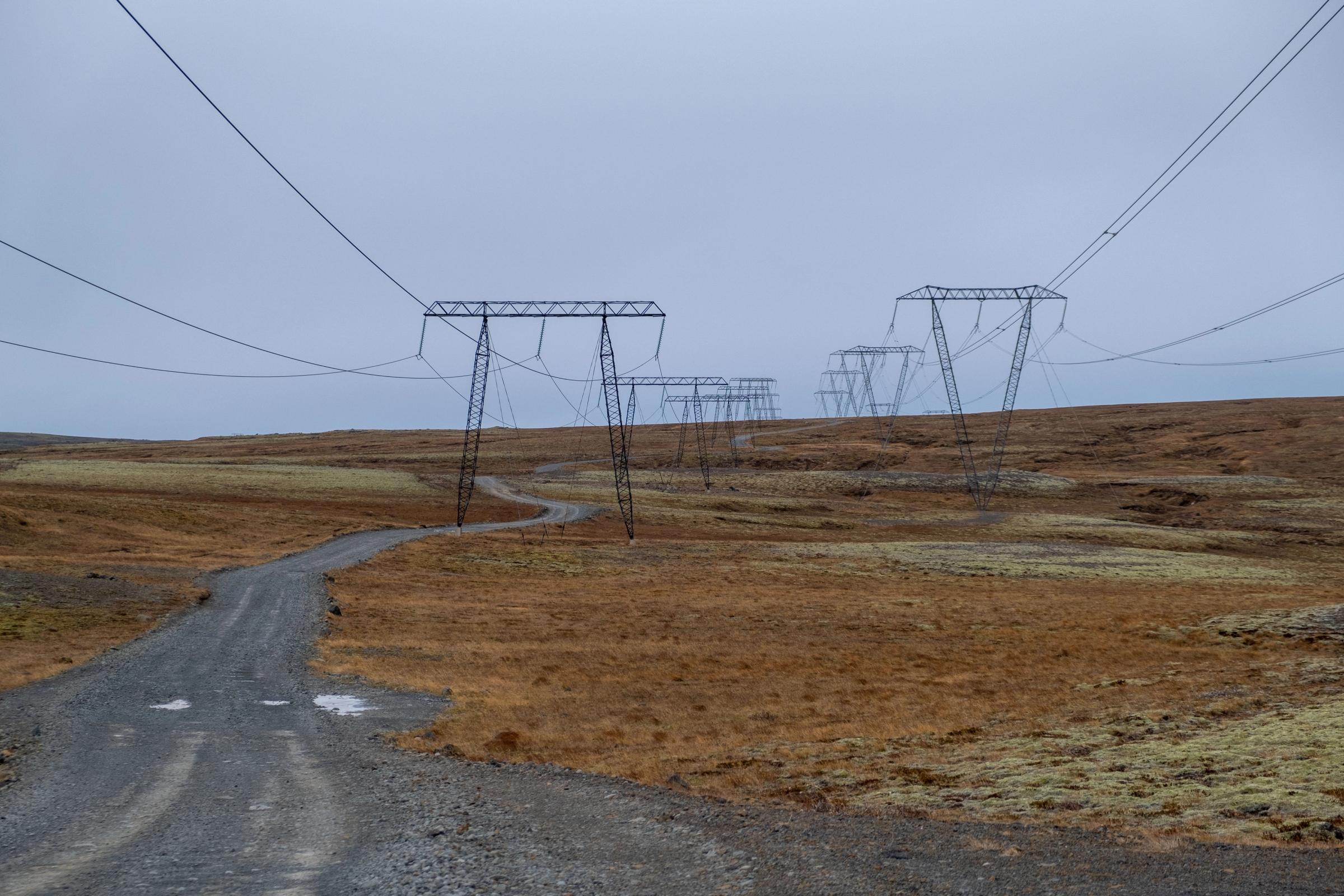 A winding gravel road leads through a dry, grassy landscape with power lines stretching into the distance under a grey sky.