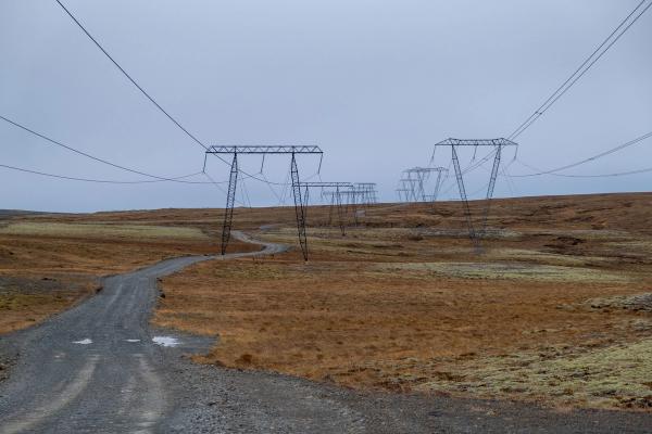 A winding gravel road leads through a dry, grassy landscape with power lines stretching into the distance under a grey sky.