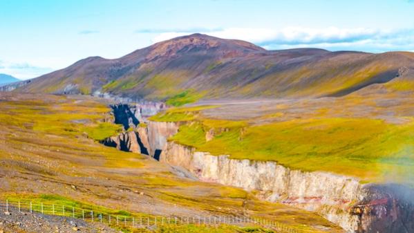 A deep canyon with colorful rock walls carves through vast yellow-green plains, with a large mountain under a blue sky.