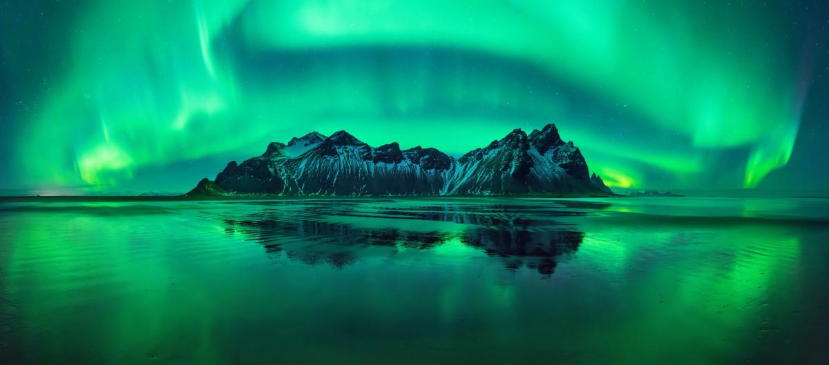 La Aurora Boreal en el cielo y reflejada en el agua al lado una montña