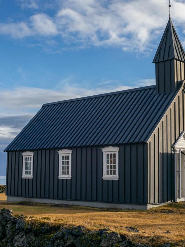 Black wooden church with a steeple on a grassy plain by the sea under a blue, cloudy sky.