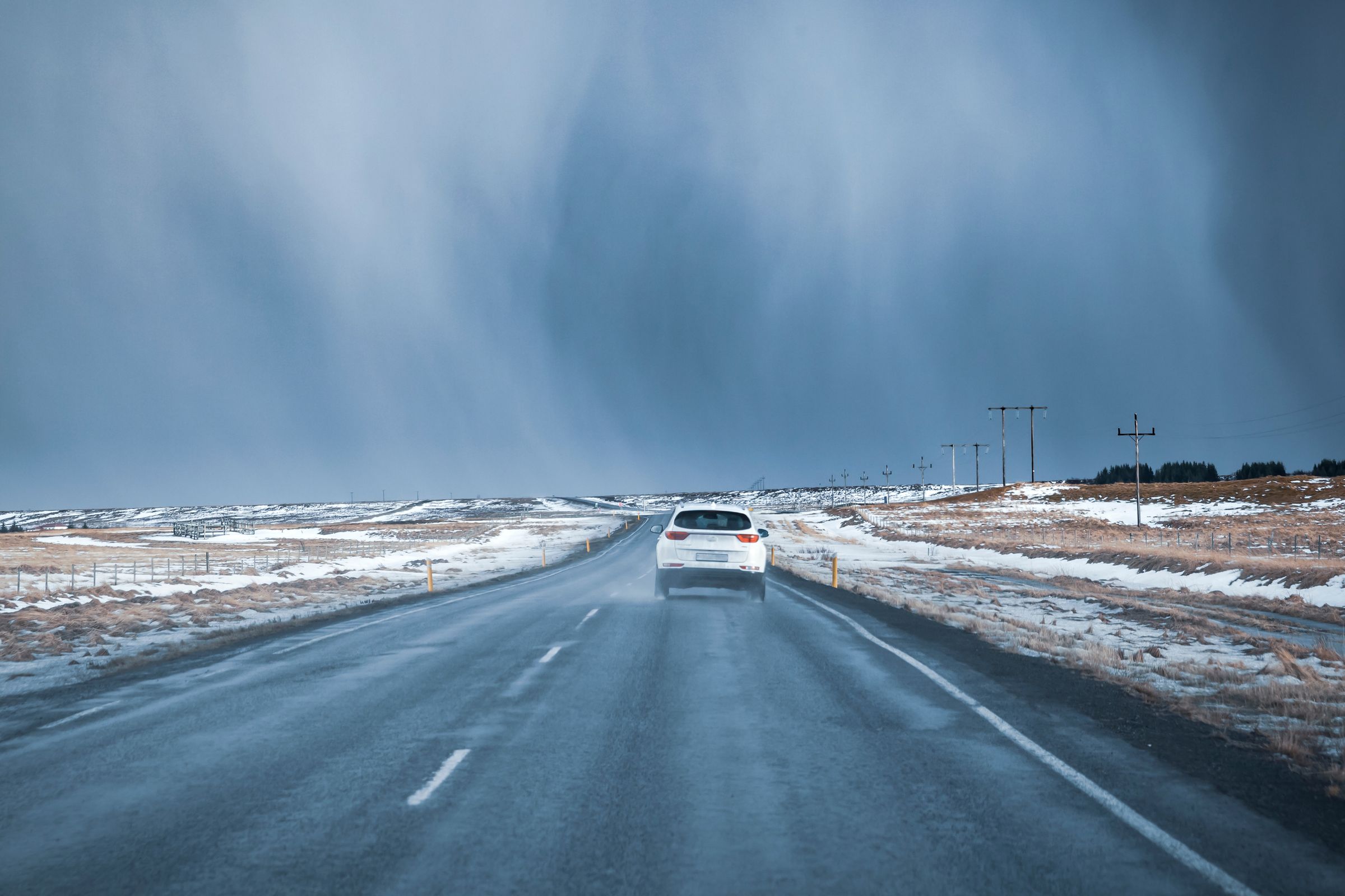 A white car drives away on a wet road through a snowy landscape under a stormy sky.