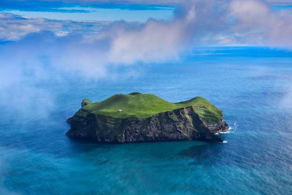 Isolated green island with a small white house and steep cliffs, surrounded by turquoise water under a cloudy sky.