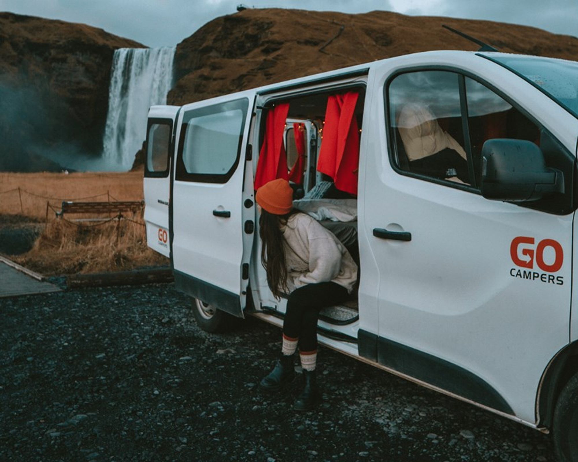a woman is getting out of a white van in front of a waterfall at a campsite, in iceland.