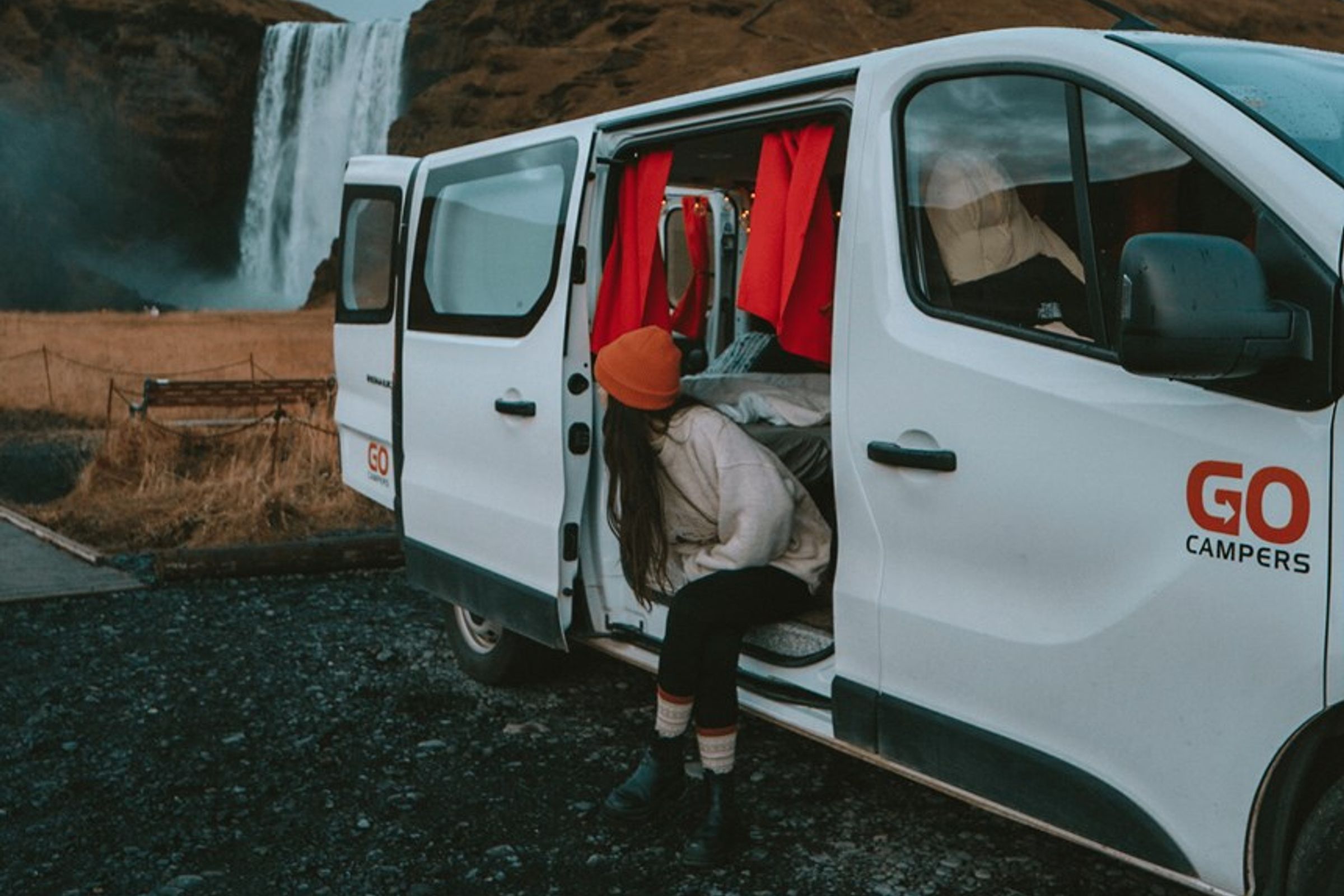 a woman is getting out of a white van in front of a waterfall at a campsite, in iceland.