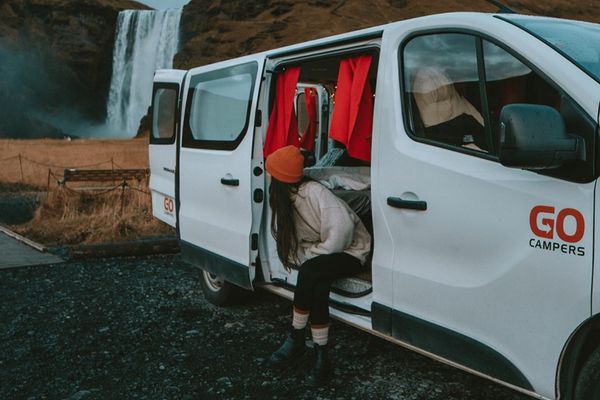 a woman is getting out of a white van in front of a waterfall at a campsite, in iceland.