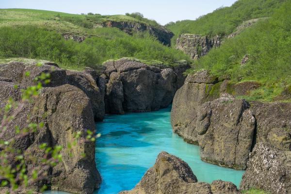 A bright turquoise river flows through a dark, rocky canyon surrounded by vibrant green hills.