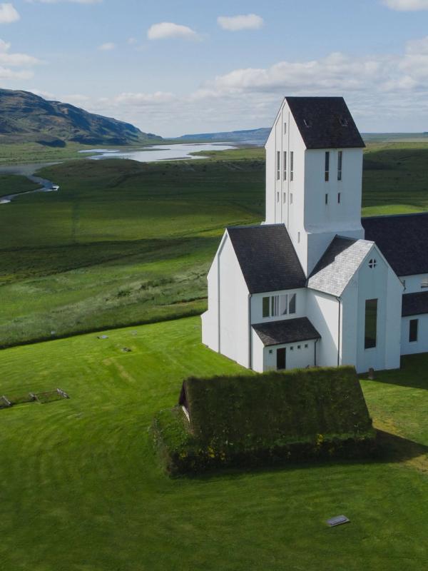 A white church with a tall tower and a small turf-roofed building in a vast green landscape with a mountain and lake in the background.