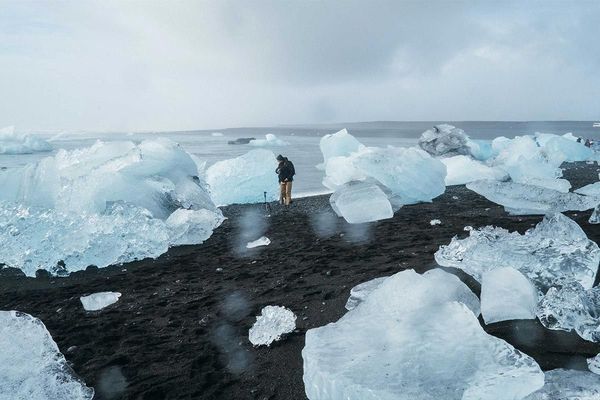 A photographer with a tripod stands on a black volcanic beach scattered with icebergs, with the ocean and a cloudy sky in the background.