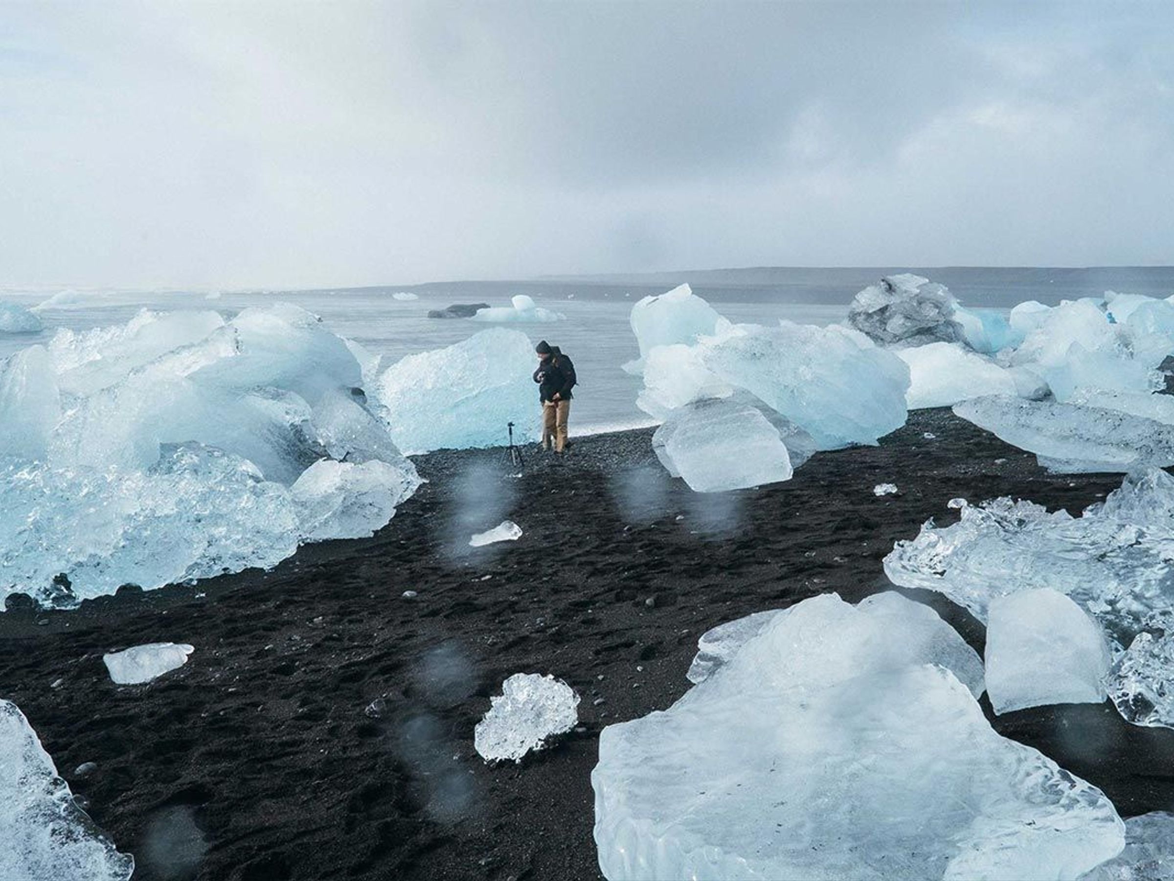 A person with a tripod on a black sand beach covered in large chunks of ice under an overcast sky.