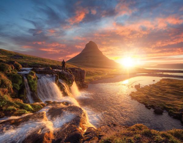 a man is standing on a rock overlooking a waterfall and a mountain at sunset .