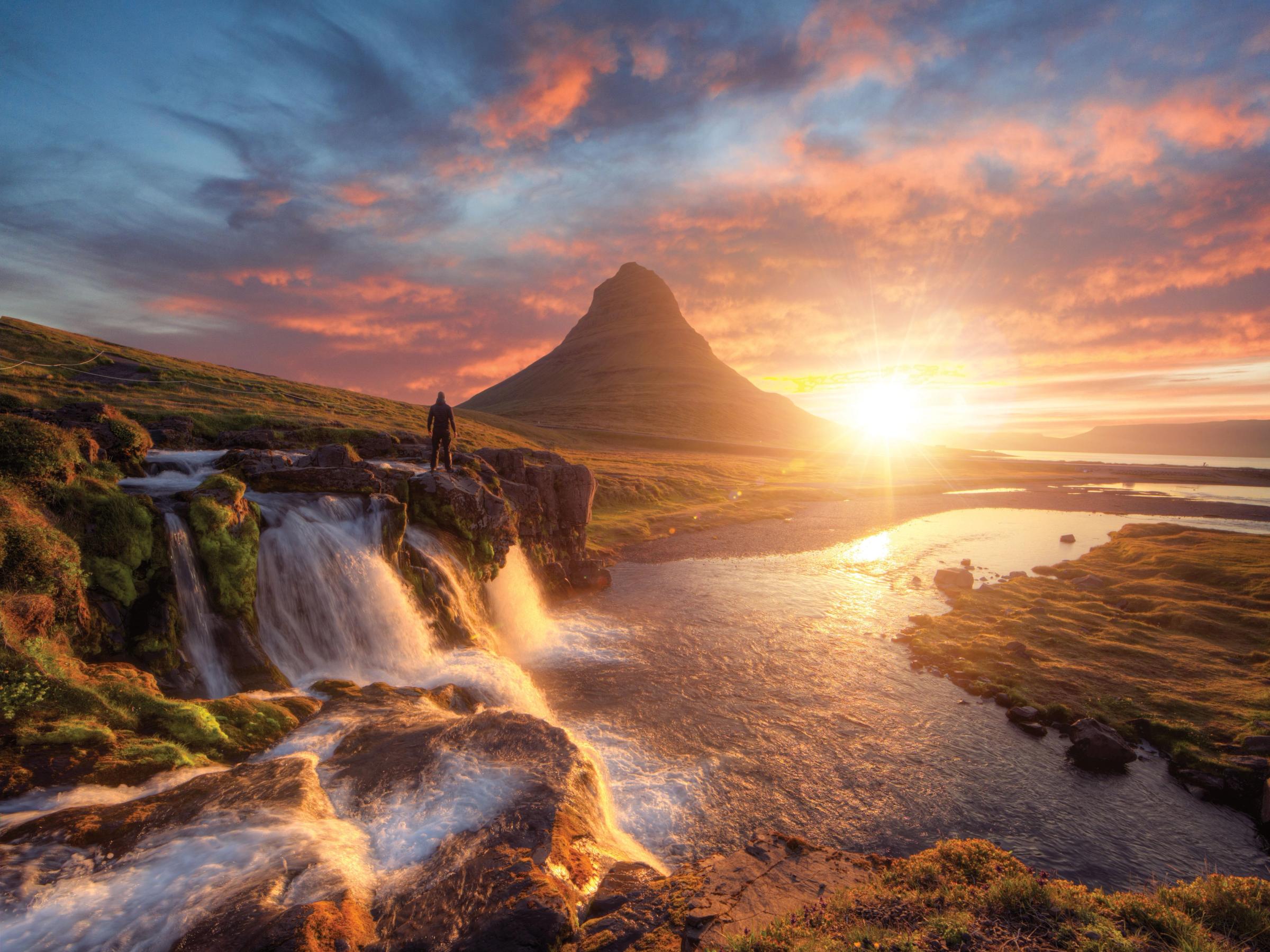 a man is standing on a rock overlooking a waterfall and a mountain at sunset .