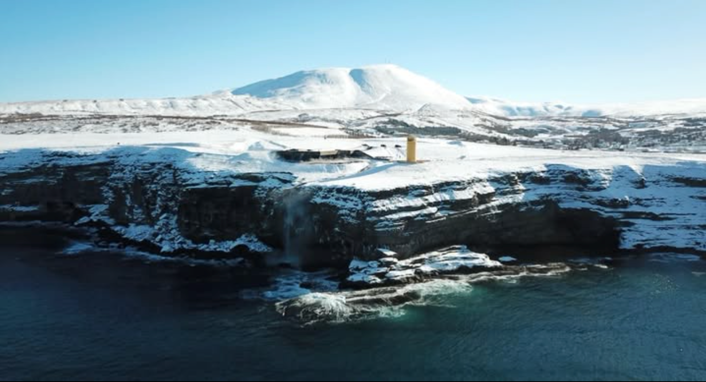 Snowy cliffs with a waterfall flowing into the ocean, a yellow lighthouse, and distant snow-capped mountains.