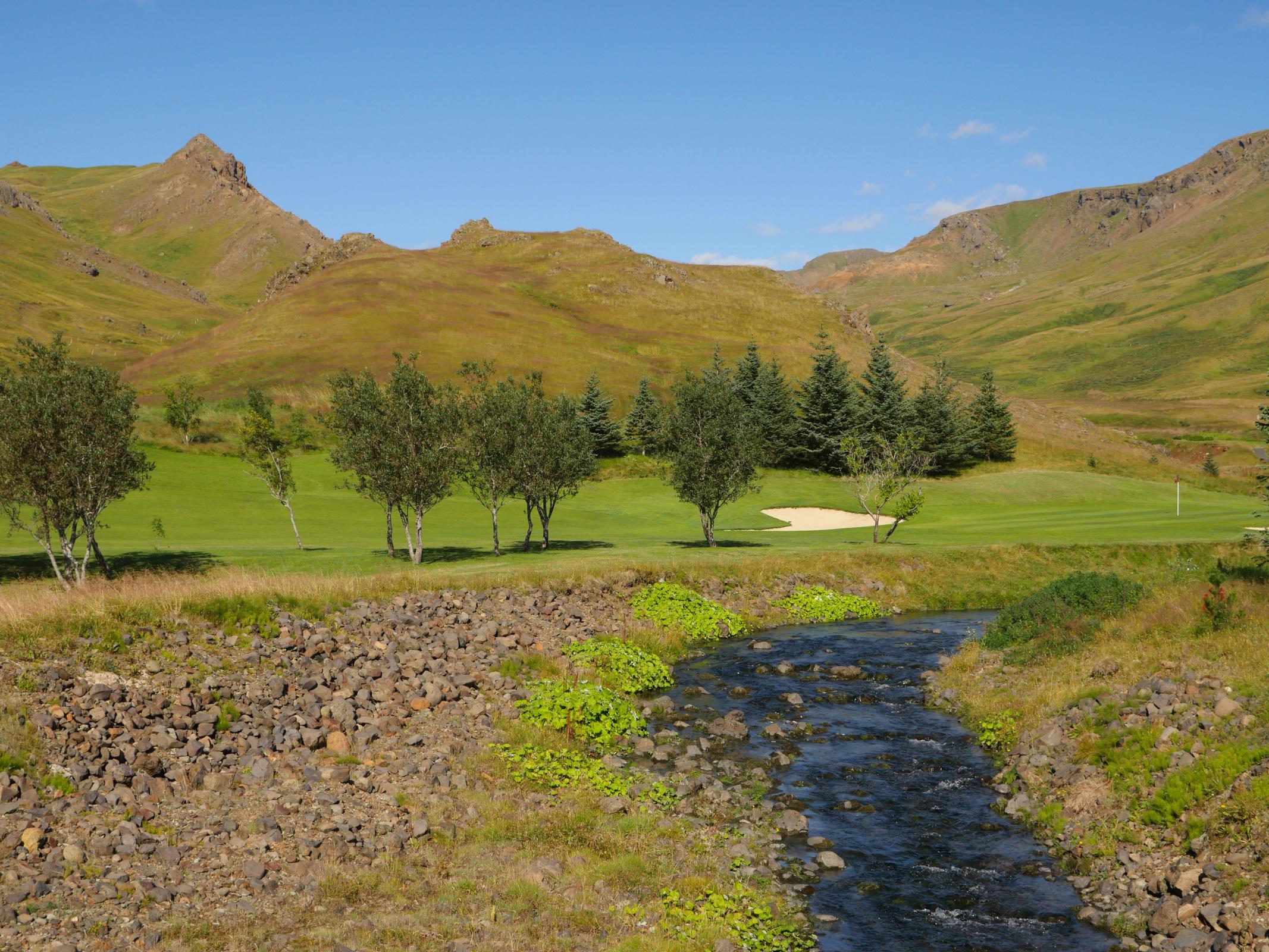 A golf course with a stream in the foreground, trees, and rolling hills under a clear blue sky.