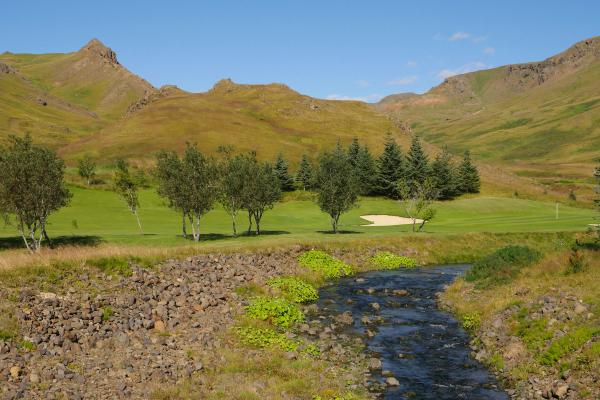 A golf course with a stream in the foreground, trees, and rolling hills under a clear blue sky.