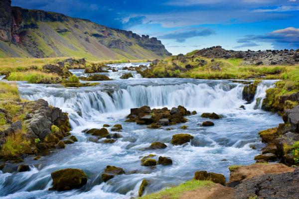 River with multiple waterfalls flowing over dark rocks, green banks, and distant mountains under a blue sky.