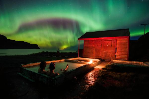 Dos personas se relajan en una terma al aire libre bajo una vibrante aurora boreal verde y morada durante la noche.