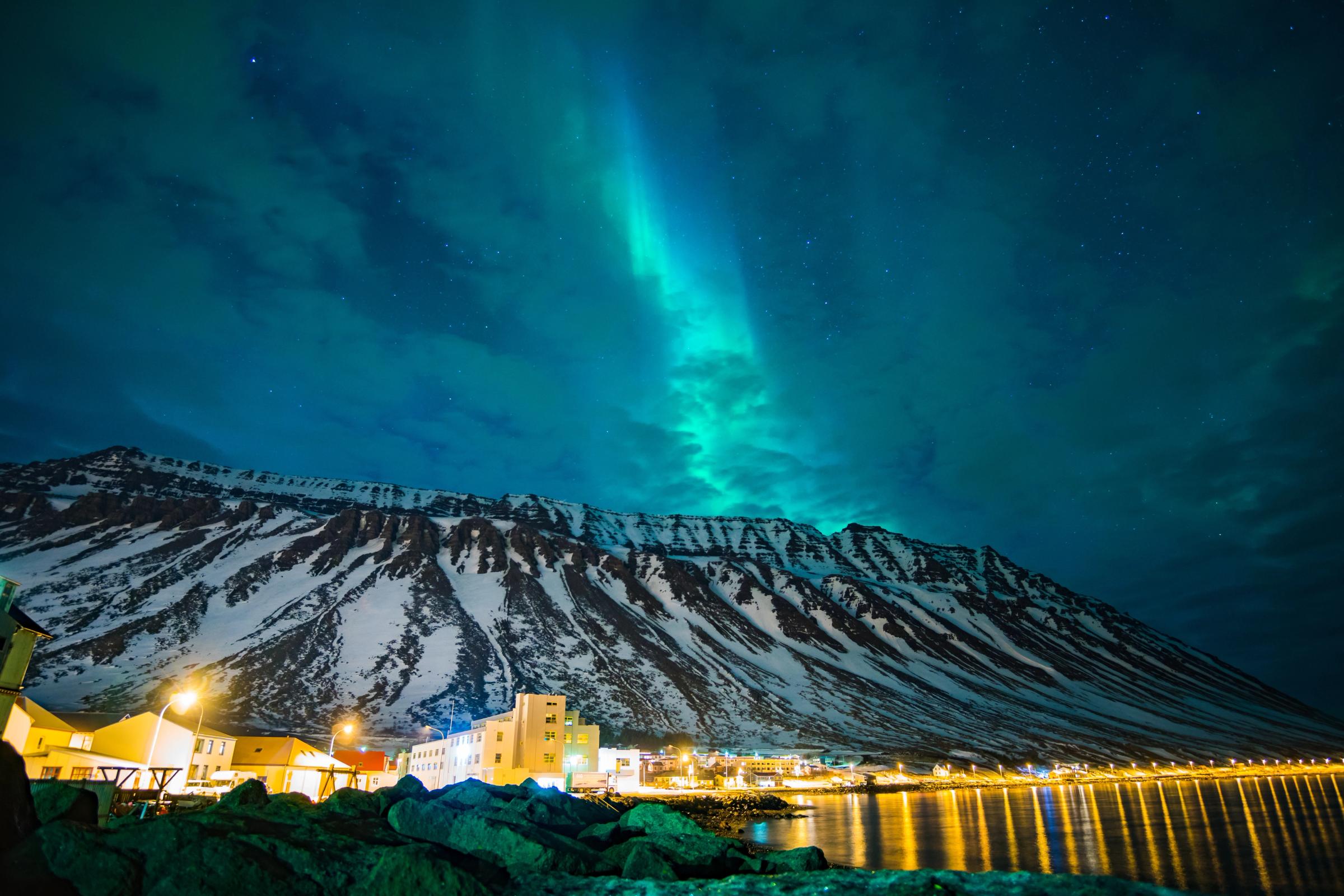 Green aurora borealis over snow-capped mountains and a lit town by a fjord.