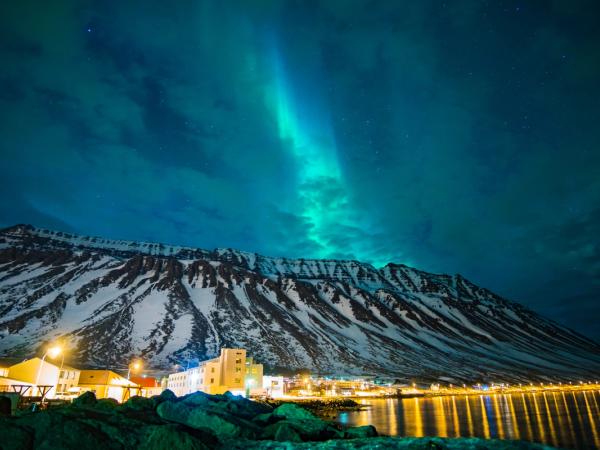 Green Northern Lights illuminate a snowy mountain range and a bright waterfront town reflecting on water at night.