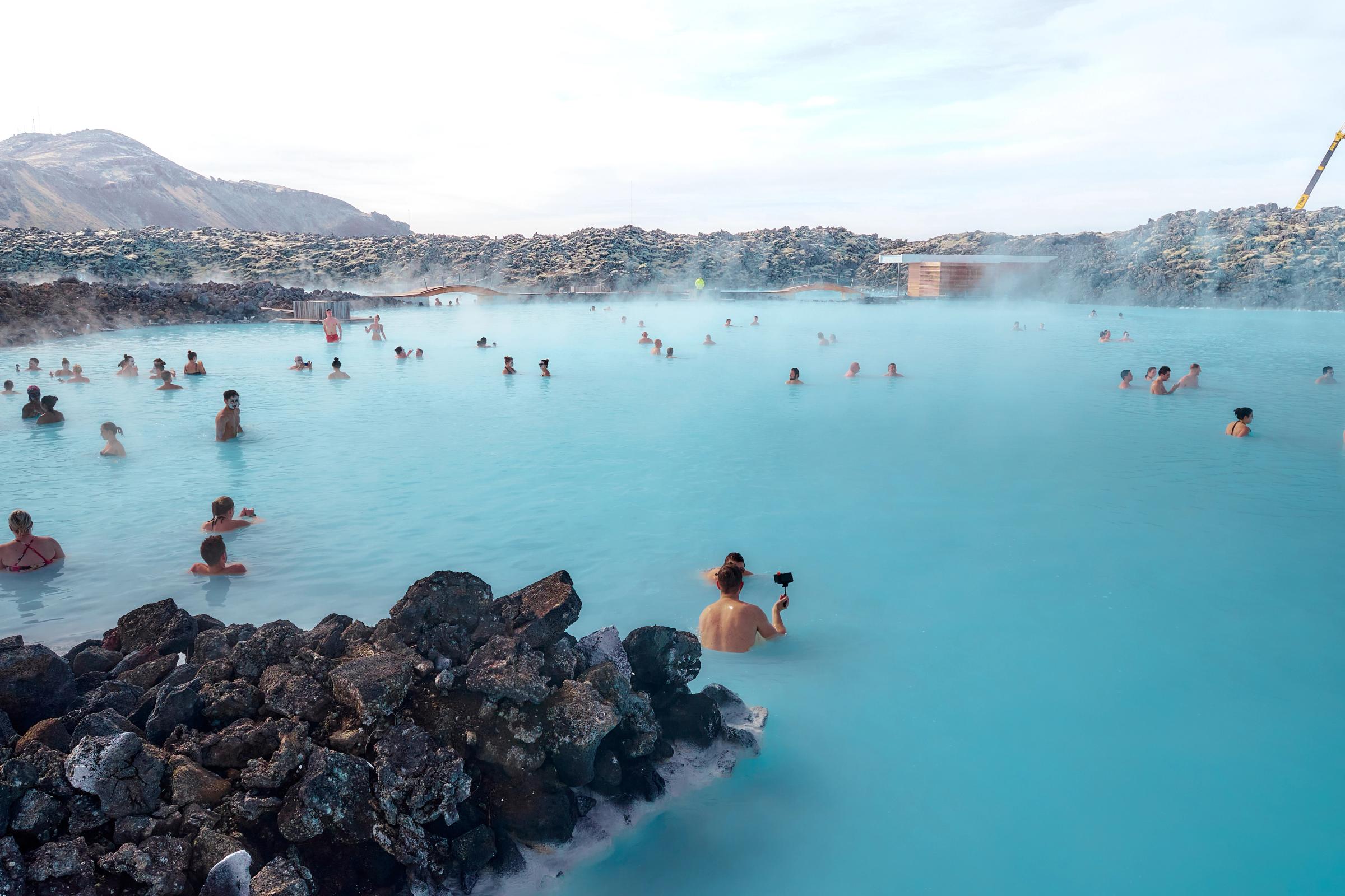 Many people bathing in a large, steaming, bright blue geothermal pool surrounded by dark volcanic rocks.