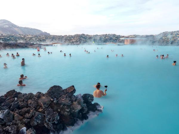 Many people bathing in a bright blue geothermal pool, surrounded by dark volcanic rocks and light steam.