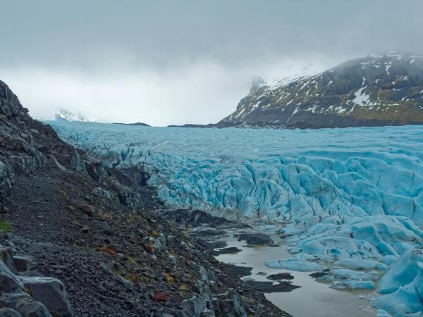 Blue glacier alongside dark rocky terrain and distant snowy mountains under an overcast sky.
