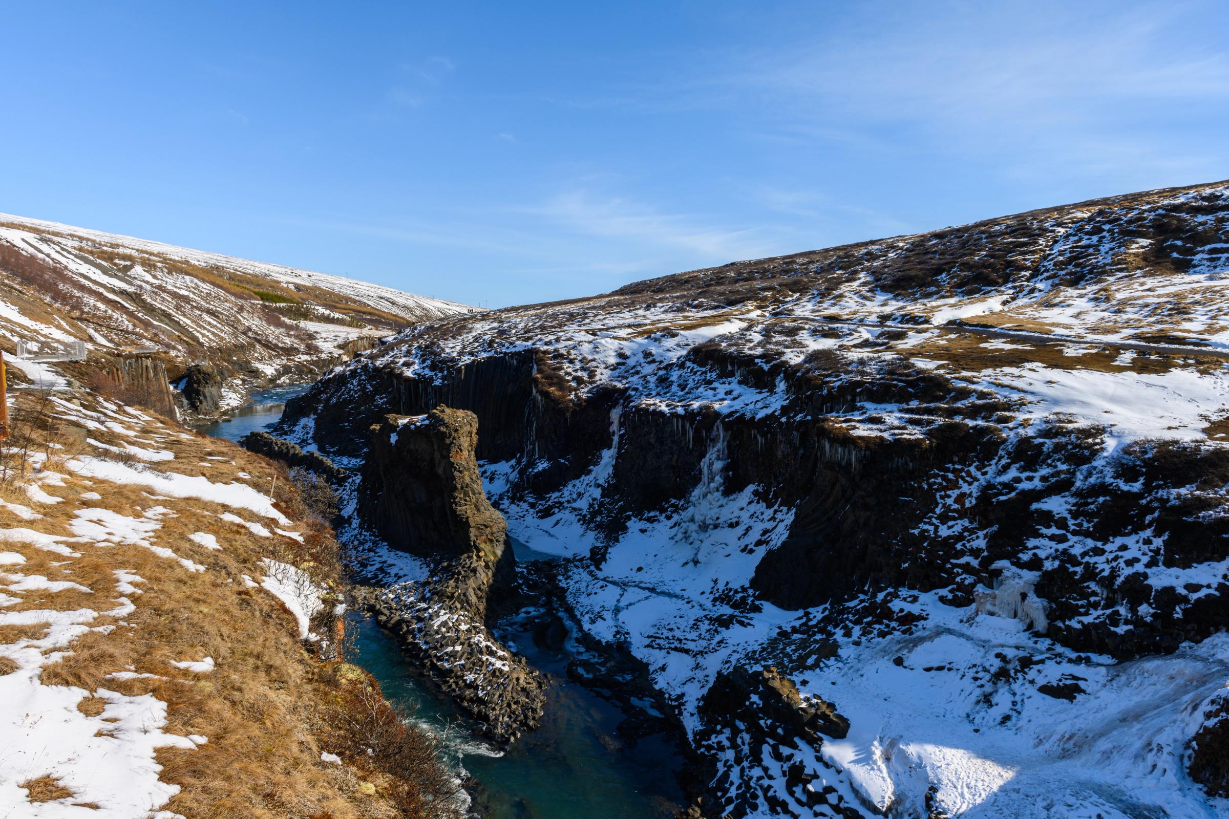 A turquoise river flows through a snow-covered canyon with rocky cliffs under a clear blue sky.
