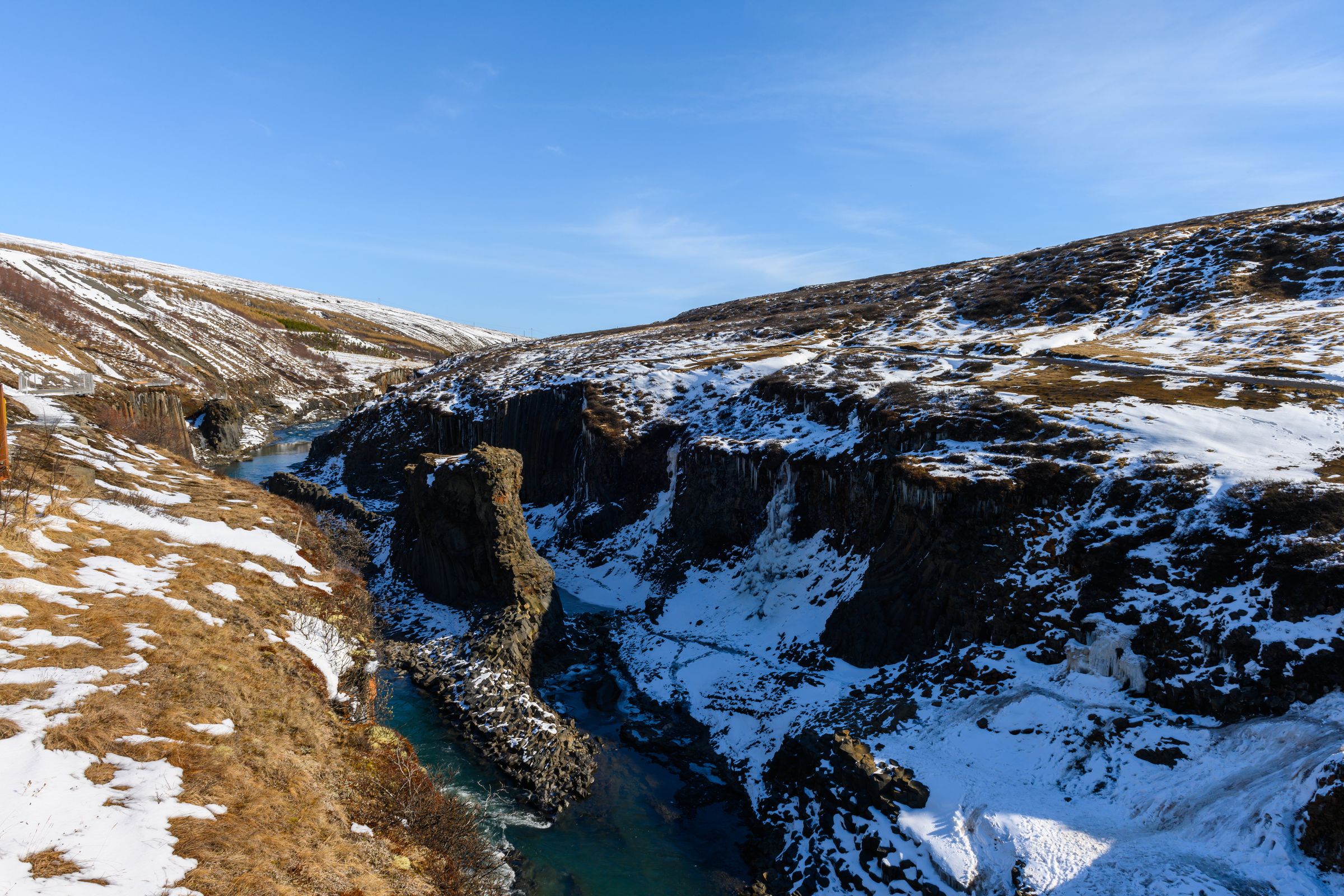 A turquoise river flows through a snow-covered canyon with rocky cliffs under a clear blue sky.