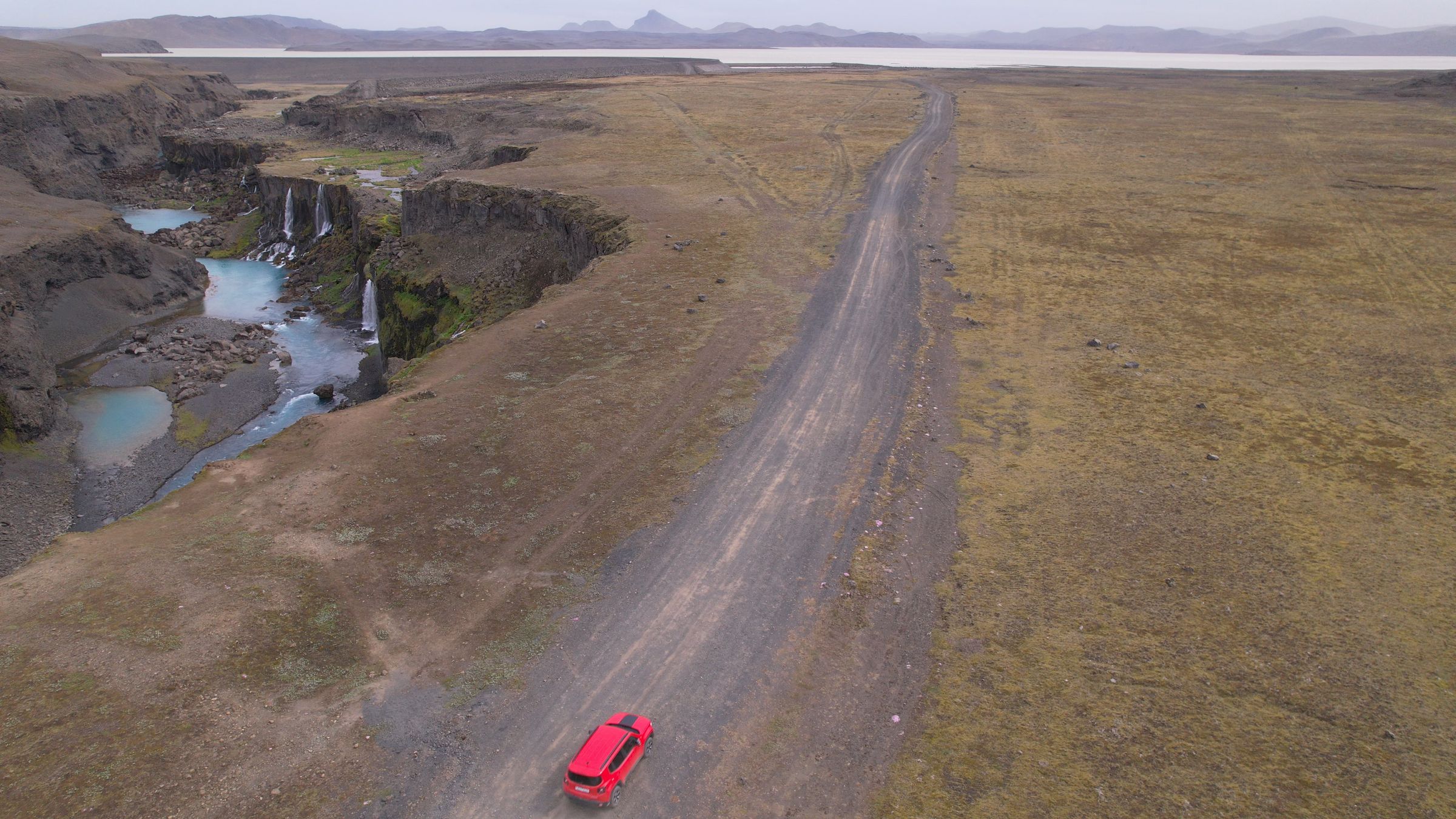 Vista aérea de un coche rojo en un camino de tierra junto a un cañón con cascadas y pozas de agua turquesa.