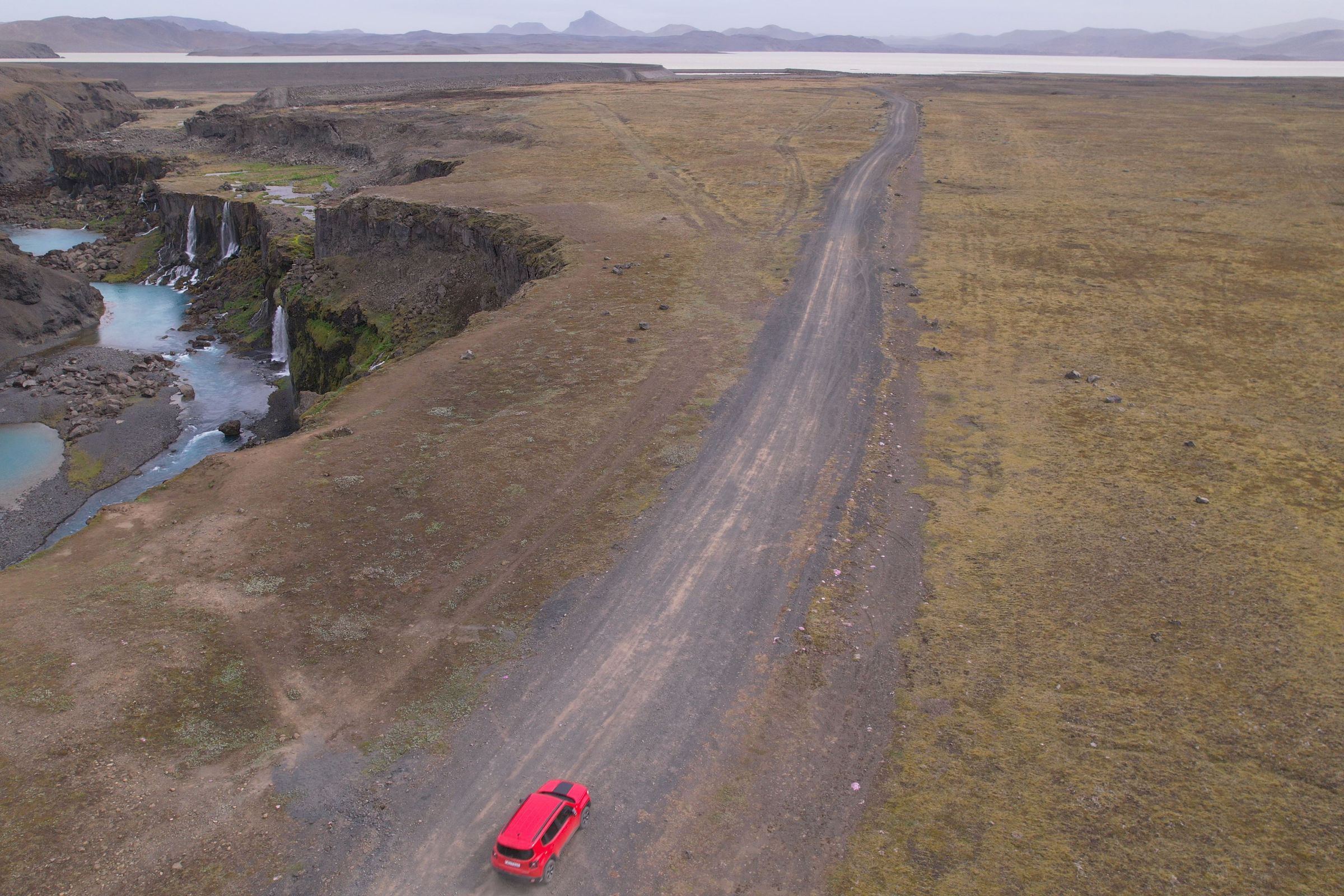 Vista aérea de un coche rojo en un camino de tierra junto a un cañón con cascadas y pozas de agua turquesa.