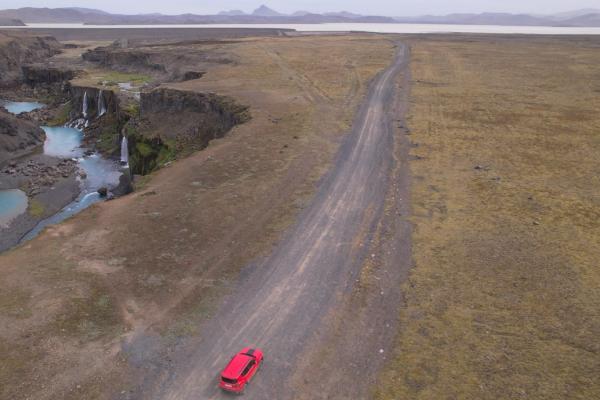Aerial view of a red car on a dirt road next to a canyon with turquoise waterfalls and pools.