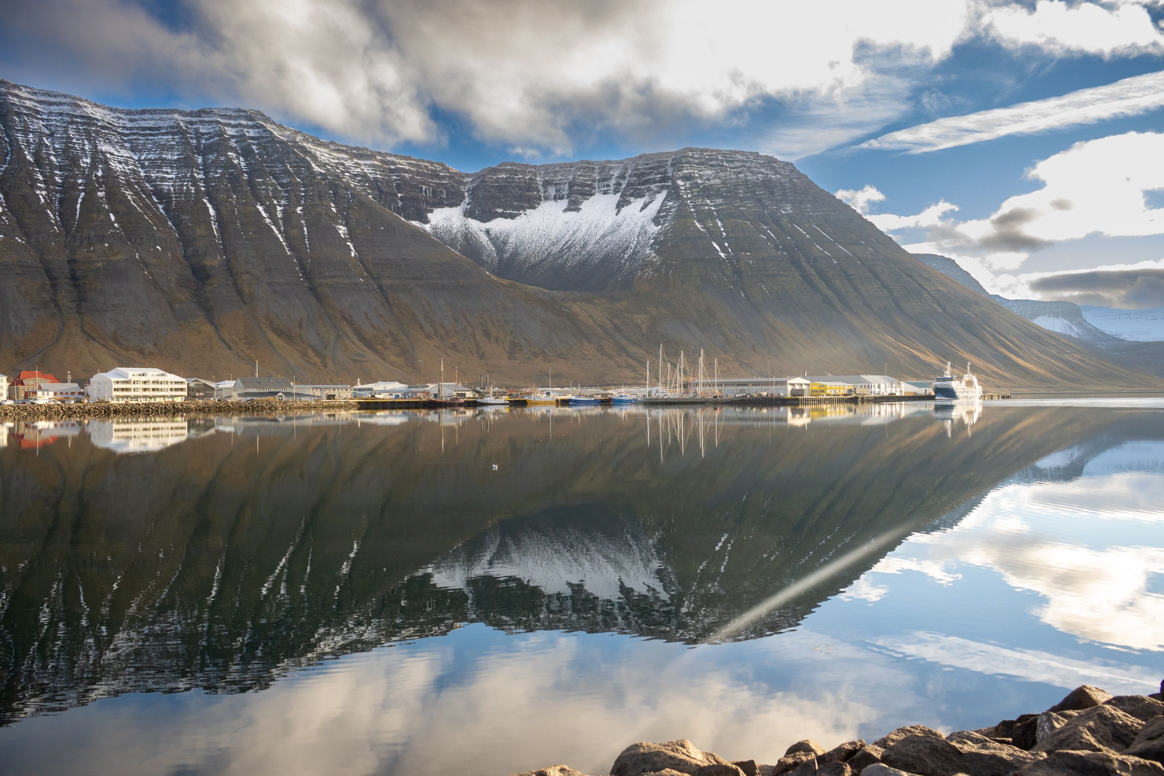 Snow-capped mountains and a village reflecting in a calm fjord.