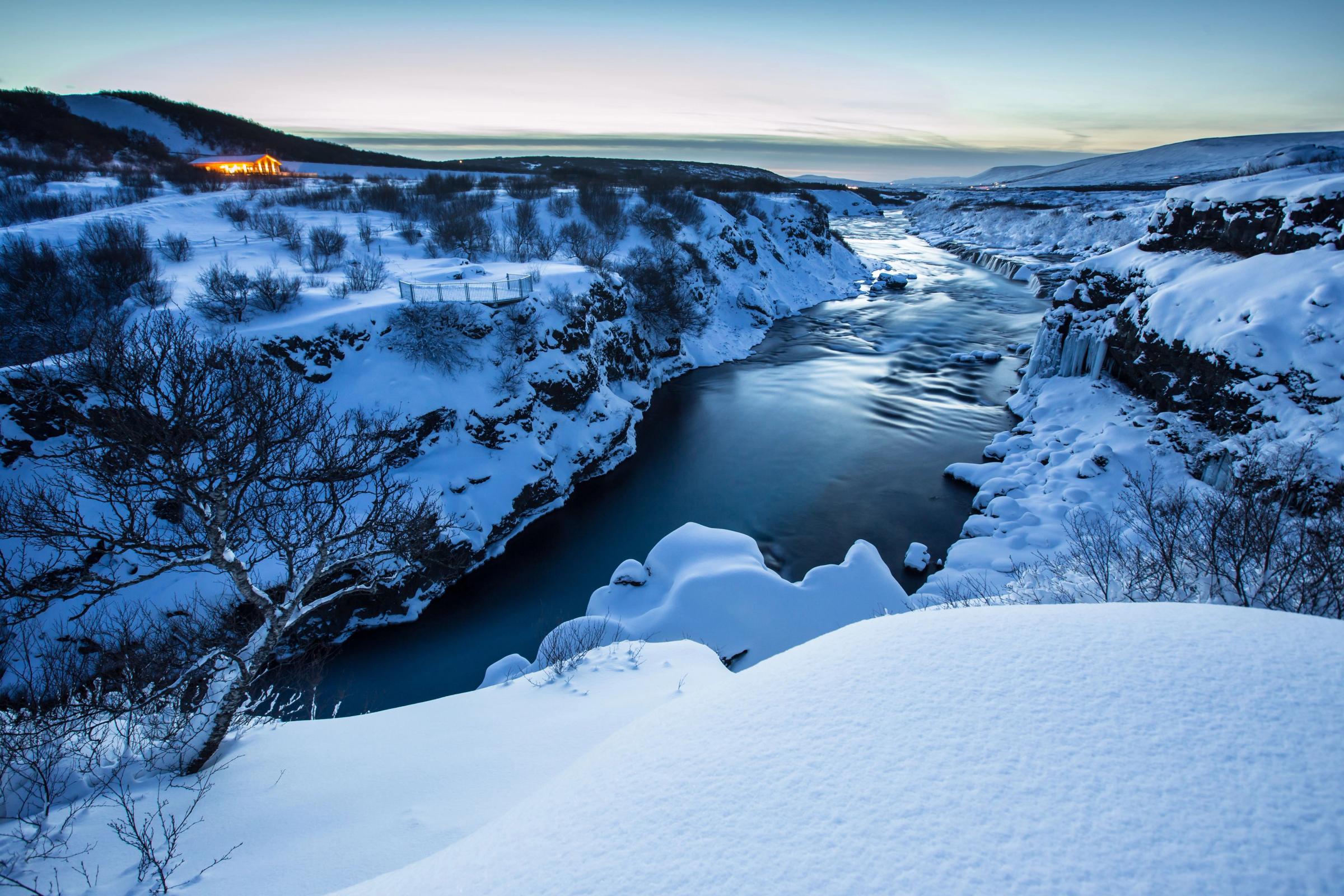 Winter scene with a blue river cutting through a snow-covered canyon and a distant lit building.