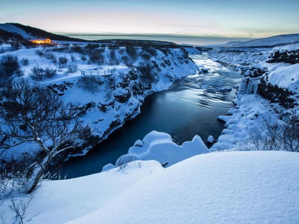 Winter scene with a blue river cutting through a snow-covered canyon and a distant lit building.