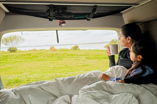A woman and child snug in a camper van bed, gazing at a green landscape and water.