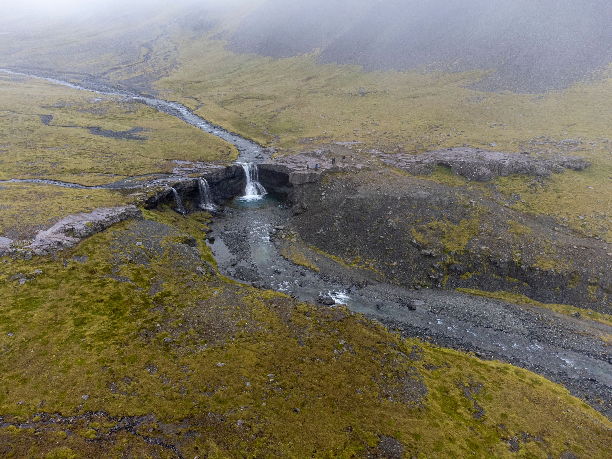 Aerial view of a river with multiple small waterfalls in a rocky, mossy landscape under a foggy sky.