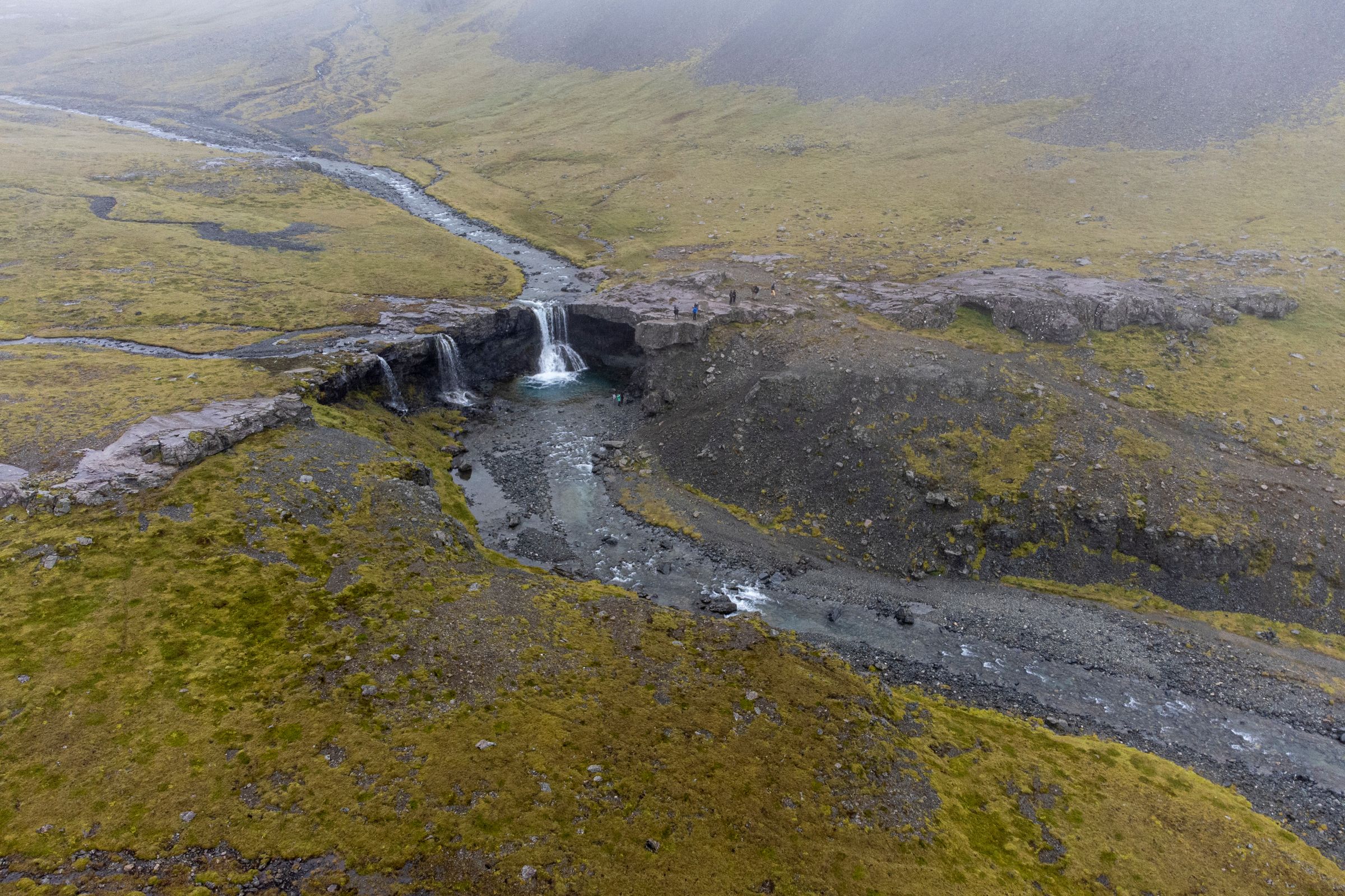Aerial view of a river with multiple small waterfalls in a rocky, mossy landscape under a foggy sky.