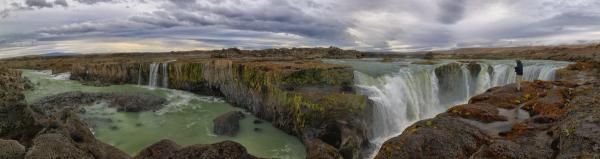 Panoramic view of a person standing by a wide, multi-tiered waterfall with green water and rocky cliffs under a cloudy sky.