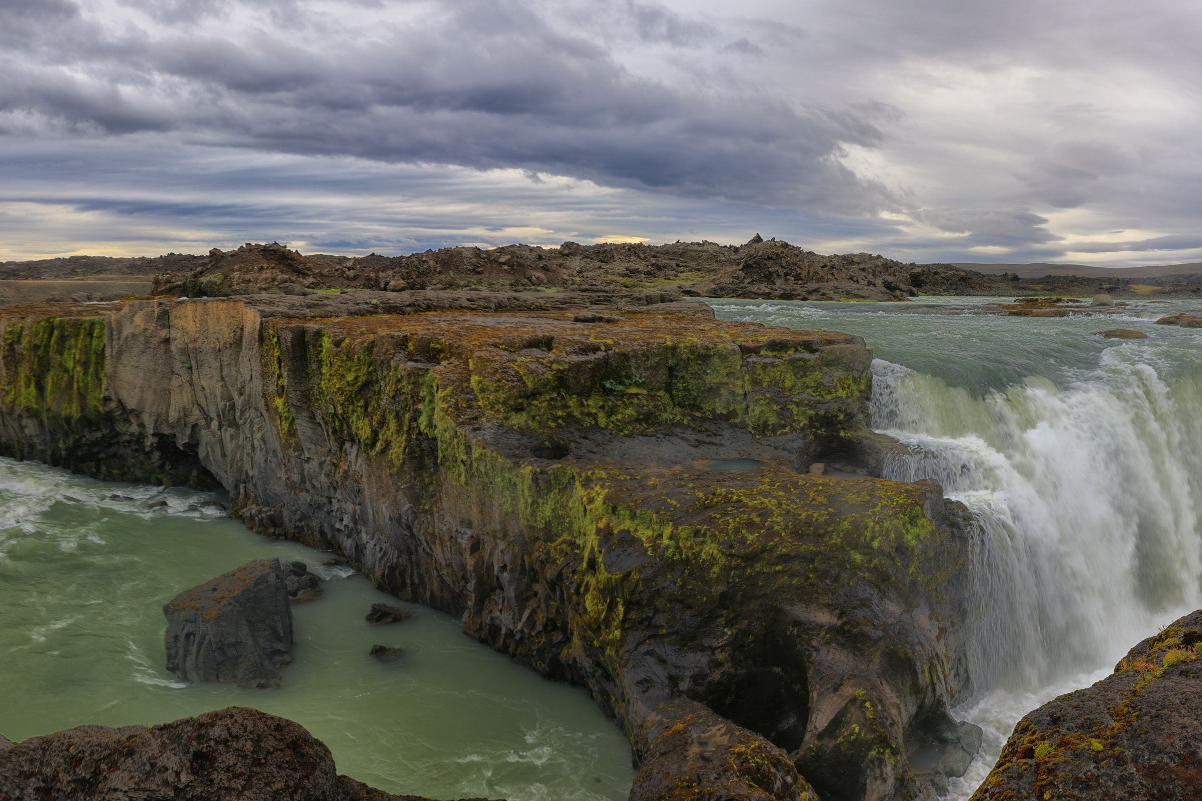 Panoramic view of a person standing by a wide, multi-tiered waterfall with green water and rocky cliffs under a cloudy sky.