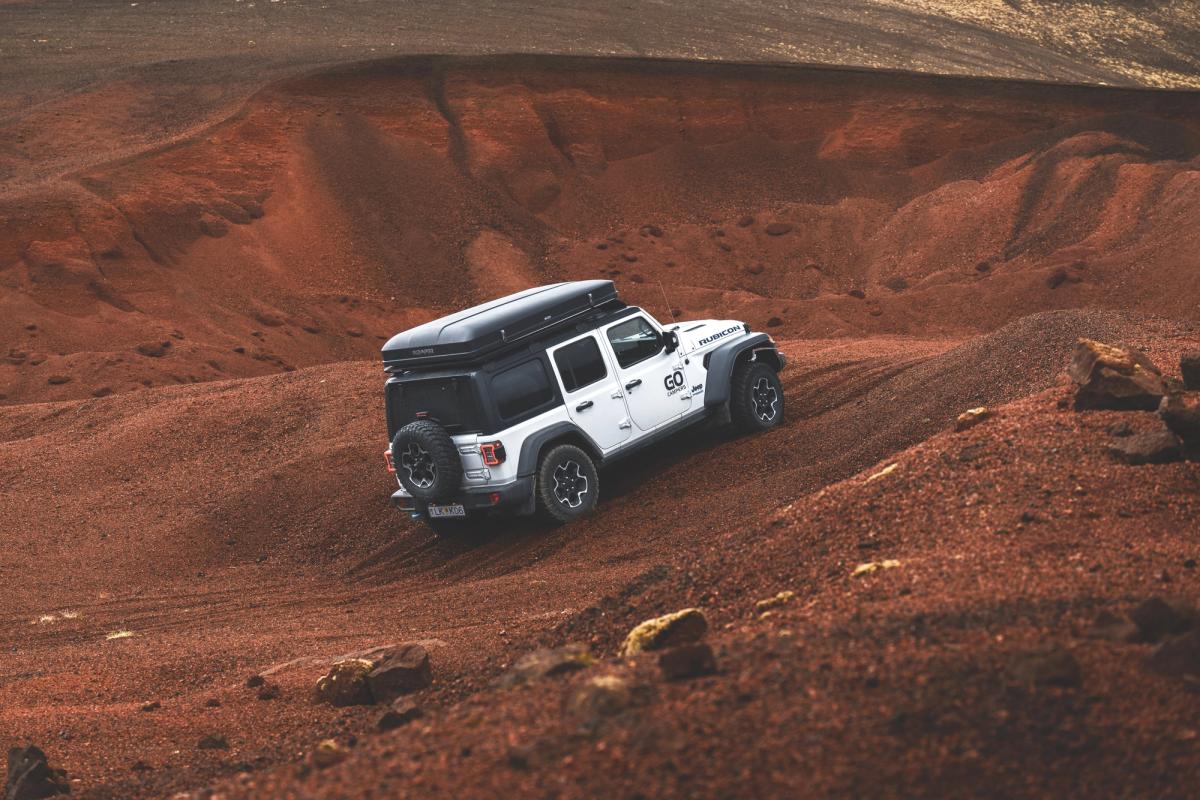 A white Jeep Wrangler with a rooftop tent drives up a steep, reddish-brown dirt hill.