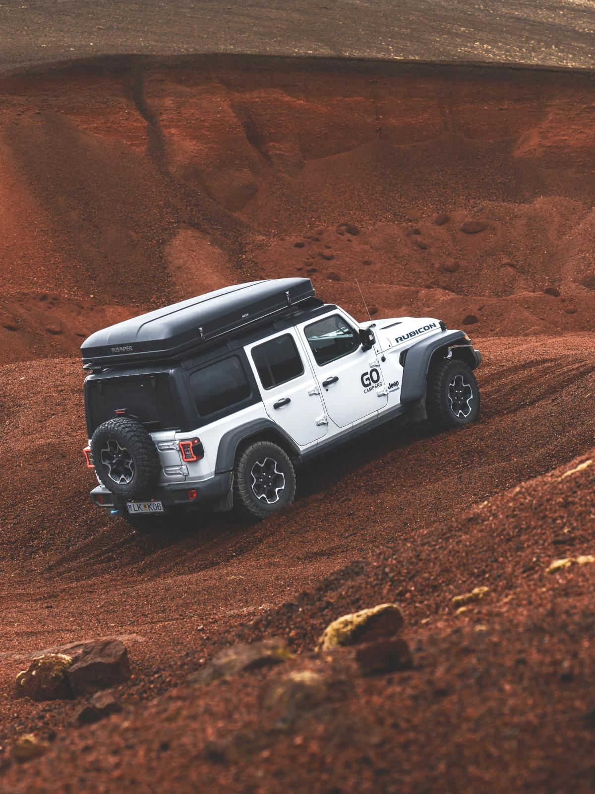 A white Jeep Wrangler with a rooftop tent drives up a steep, reddish-brown dirt hill.