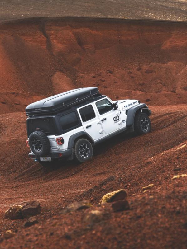 A white Jeep Wrangler with a rooftop tent drives up a steep, reddish-brown dirt hill.