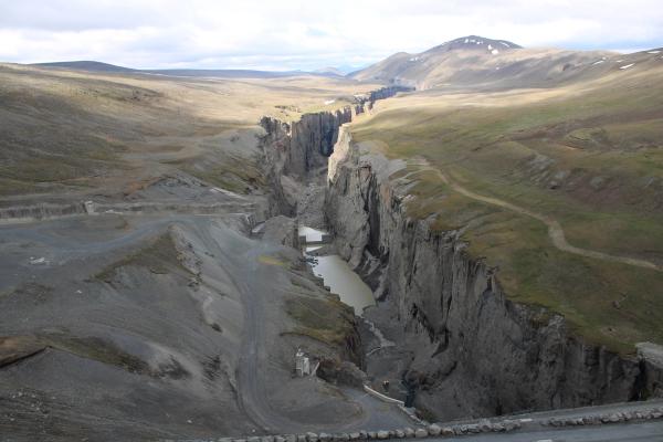 A deep, narrow canyon with steep, rocky walls and light-colored water pools, set within a vast landscape of rolling, sparsely vegetated hills and distant mountains under a cloudy sky.