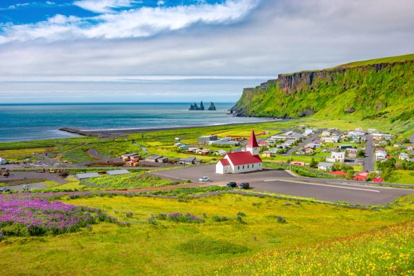 Coastal village with a red-roofed church, green cliffs, black sand beach, and sea stacks.