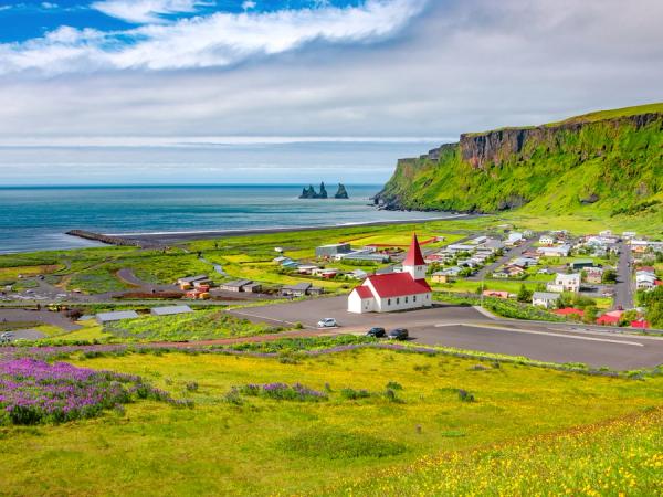 A vibrant coastal village with a white church, green cliffs, black sand beach, and distant sea stacks.