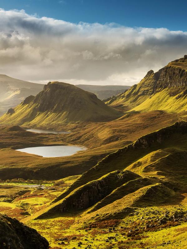 Dramatic landscape of rolling green and brown hills with two small lakes under a partly cloudy sky.