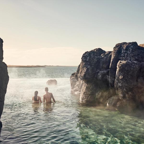 A man and woman standing in a steaming geothermal pool surrounded by dark rock formations.